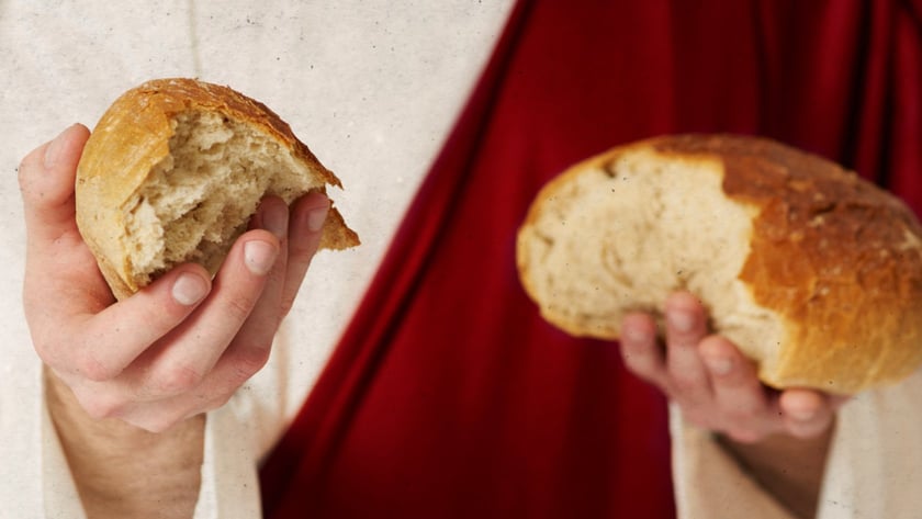 Pessoa de túnica clara e manto vermelho segura dois pedaços de pão partido.