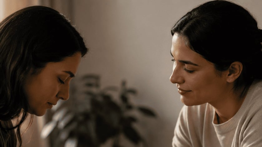 Duas mulheres sentadas de mãos dadas em uma sala, uma delas de cabeça baixa e olhos fechados.