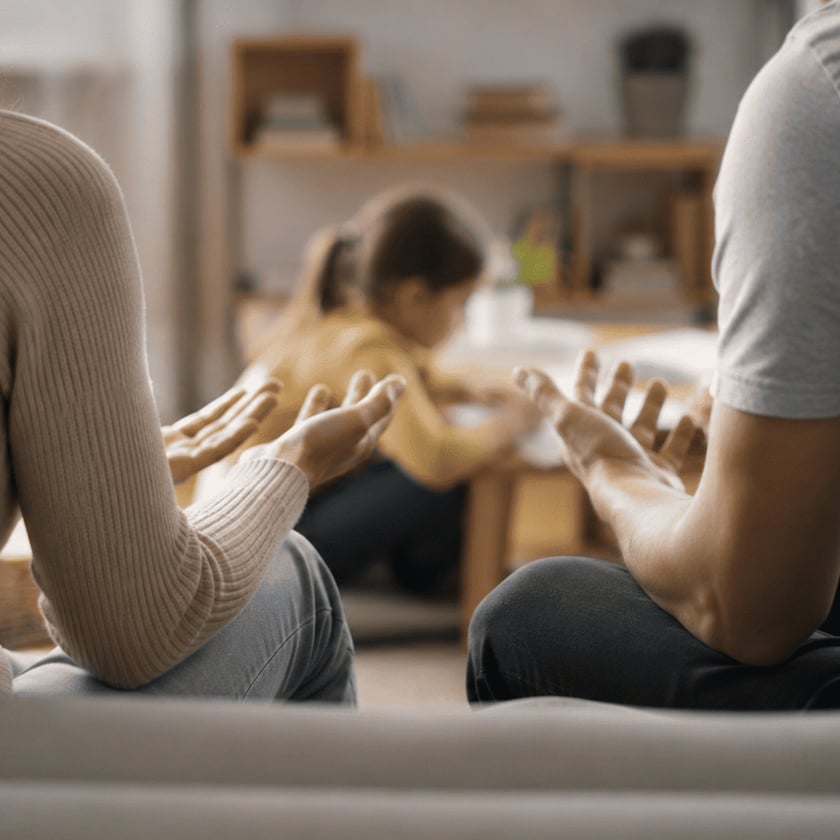 Duas pessoas sentadas num sofá com as palmas das mãos abertas; ao fundo, uma criança brinca.