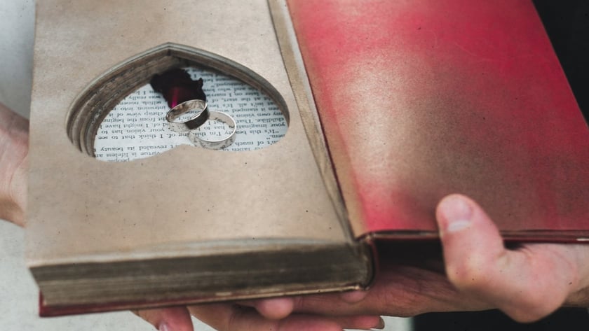 Duas pessoas seguram um livro aberto com recorte em coração contendo duas alianças de prata.