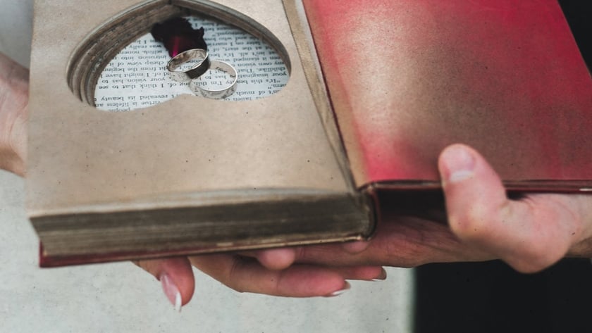 Duas pessoas seguram um livro aberto com recorte em coração contendo duas alianças de prata.