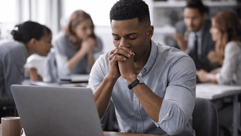 Homem de camisa azul com as mãos unidas diante do rosto, sentado diante de um notebook.