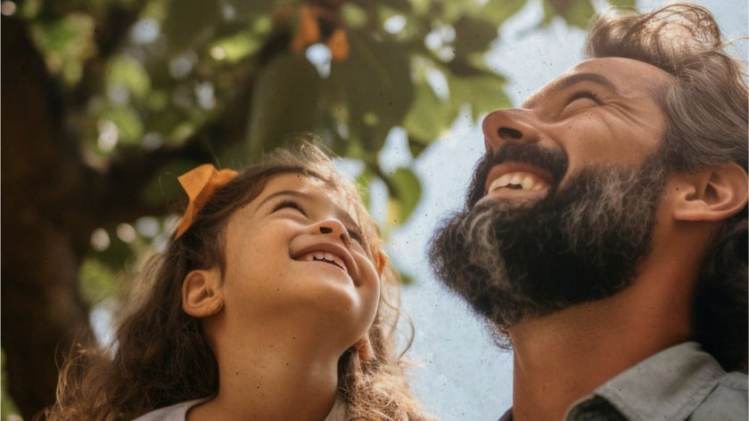 Homem barbudo e menina de cabelo cacheado olham para cima, sorrindo, sob uma árvore.