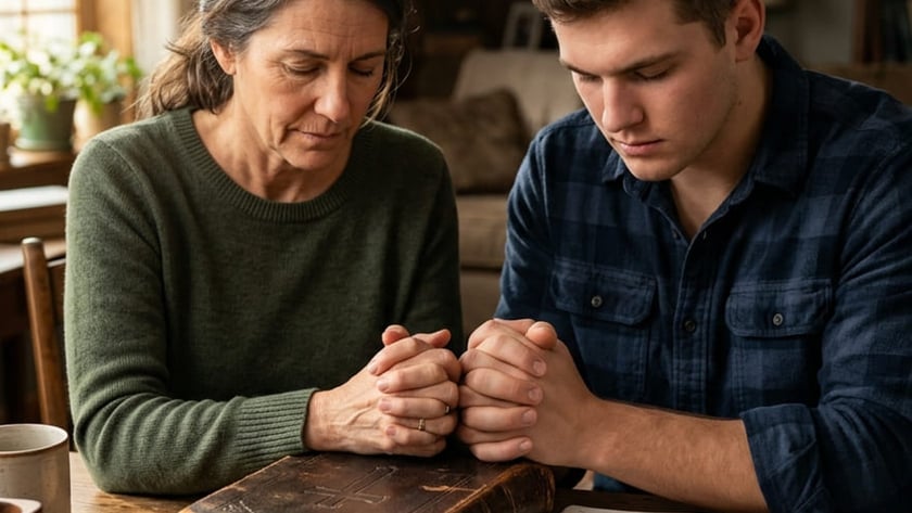 Mulher e rapaz de mãos juntas e olhos fechados, rezando à mesa sobre uma Bíblia.