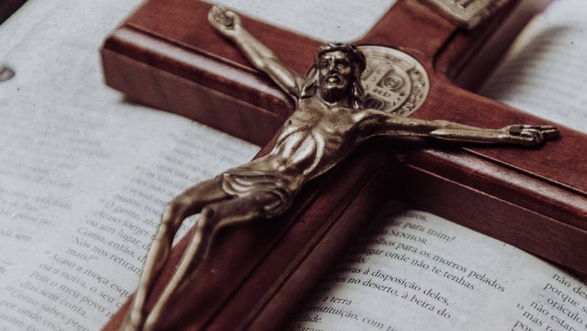 Crucifixo de madeira com Cristo metálico sobre Bíblia aberta, com tecido roxo ao fundo. "INRI"