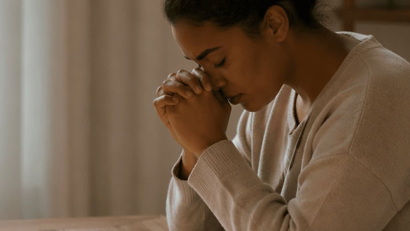 Mulher de olhos fechados e mãos postas sobre uma Bíblia aberta, à mesa, ao lado de vela e caneca.