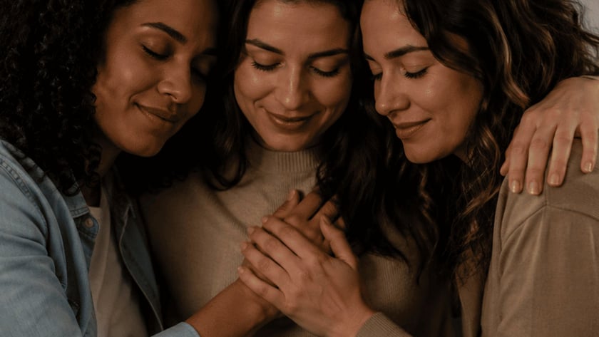 Três mulheres abraçadas com as cabeças encostadas e as mãos unidas sobre o peito da do meio.