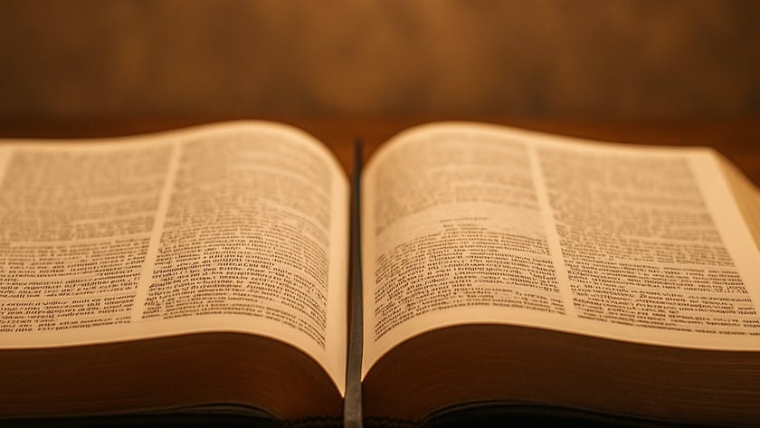 Livro grande aberto, com duas páginas de texto e uma fita marcadora, sobre mesa de madeira.