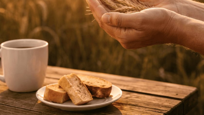 Mãos seguram espigas de trigo diante de uma xícara e torradas sobre mesa de madeira.
