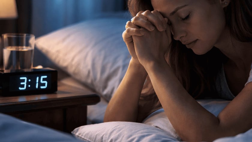 Mulher deitada na cama de mãos postas junto ao rosto, ao lado de um relógio marcando "3:15".