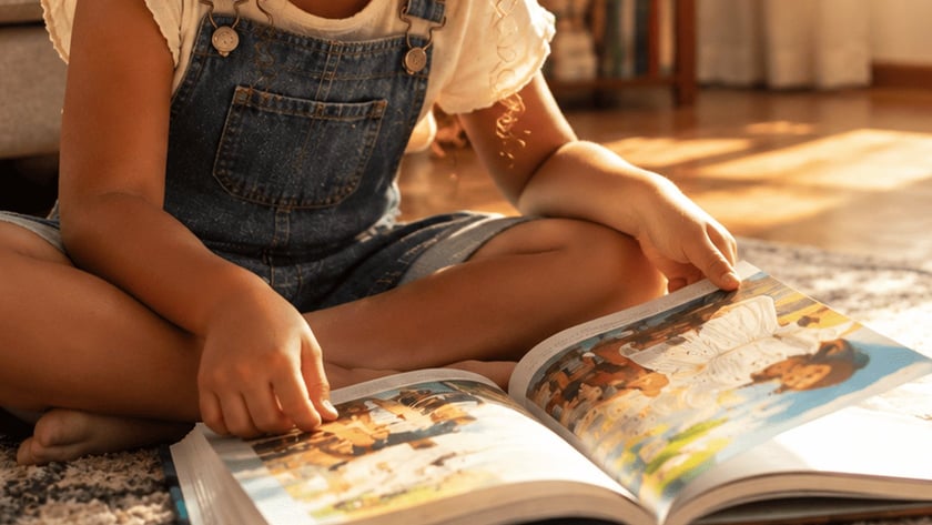 Menina de cabelo cacheado sentada no chão folheia um livro ilustrado aberto em uma sala.