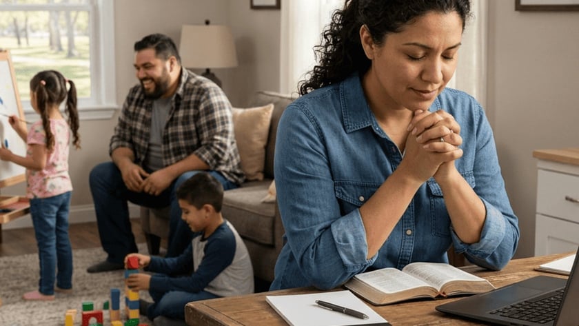 Mulher de olhos fechados e mãos juntas à mesa, com Bíblia; ao fundo, família brinca na sala.