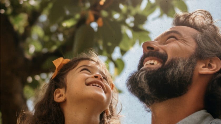 Homem barbudo e menina de cabelo cacheado olhando para cima, sob uma árvore.