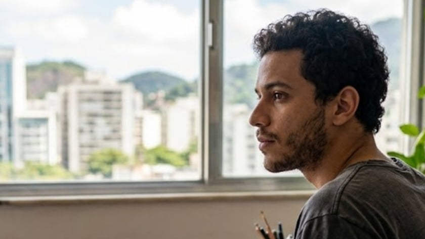 Homem de cabelo cacheado desenha em caderno ao lado de uma janela com prédios.
