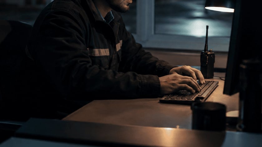 Homem de jaqueta digita no computador, em uma mesa ao lado de um rádio, à noite.