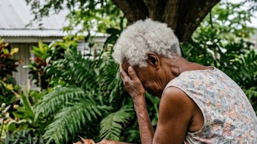 Idosa de cabelo branco sentada num banco no jardim, com a mão no rosto, segurando um terço.