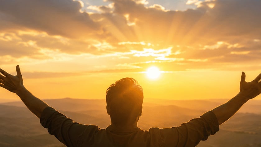 Homem de costas, com os braços abertos, olha o pôr do sol sobre colinas.