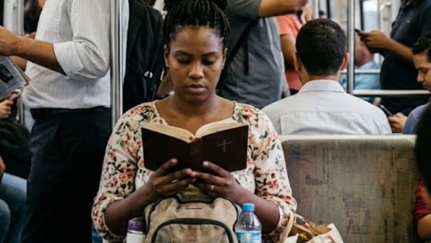 Mulher sentada em vagão de metrô lotado lê uma Bíblia, com mochila no colo.