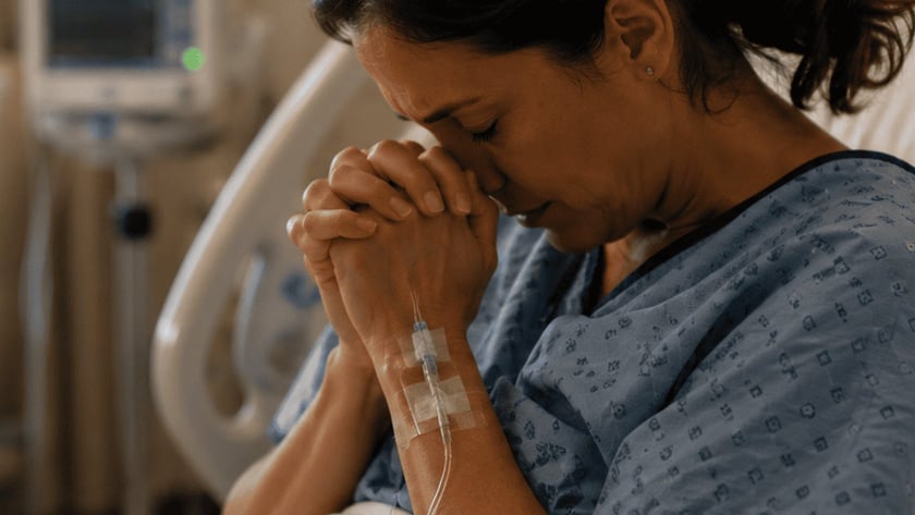 Mulher de avental hospitalar, sentada na cama, com as mãos unidas junto ao rosto e soro no braço.