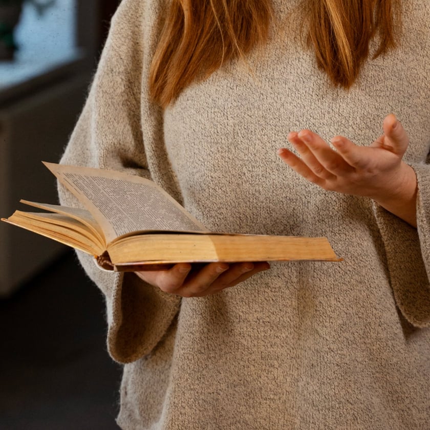 Mulher de cabelo ruivo e suéter bege segura um livro antigo aberto e gesticula.