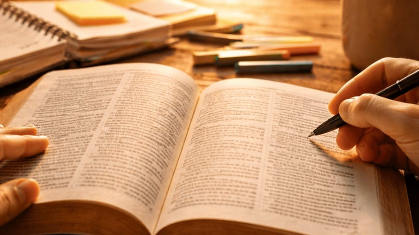 Mãos de uma pessoa sobre um livro aberto, com caneta, cadernos e xícara na mesa.