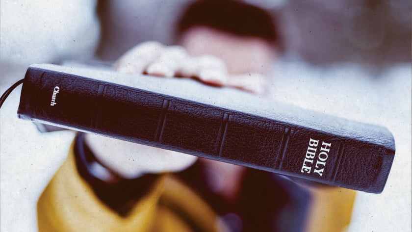 Pessoa de casaco amarelo estende uma Bíblia preta de capa de couro: "HOLY BIBLE".