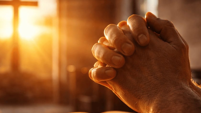 Mãos postas ao lado de uma Bíblia aberta, diante de uma cruz contra a janela.