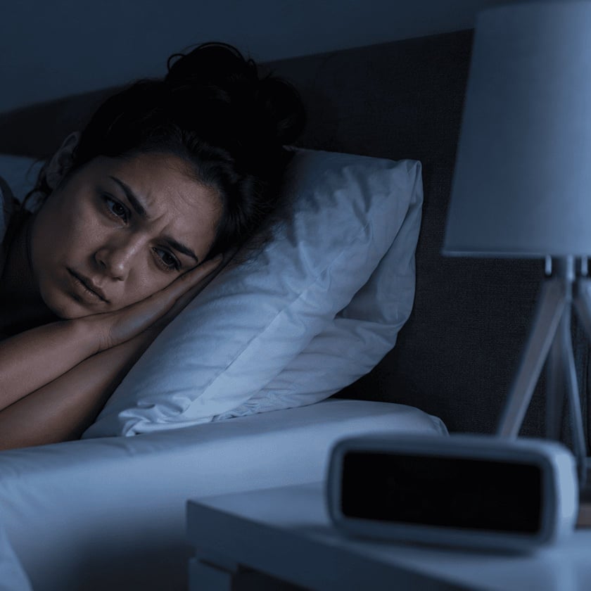 Mulher deitada na cama, de olhos abertos, olhando para o despertador na mesa de cabeceira.