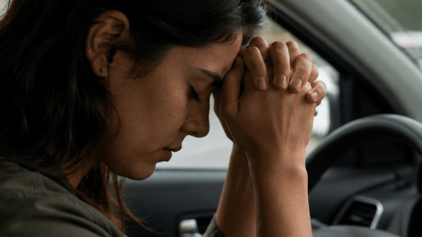 Mulher ao volante de um carro com a testa apoiada nas mãos entrelaçadas, em trânsito parado.