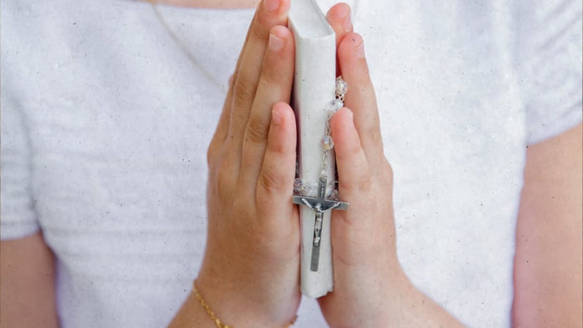 Criança de blusa branca segura as mãos postas com um livro branco e um terço.