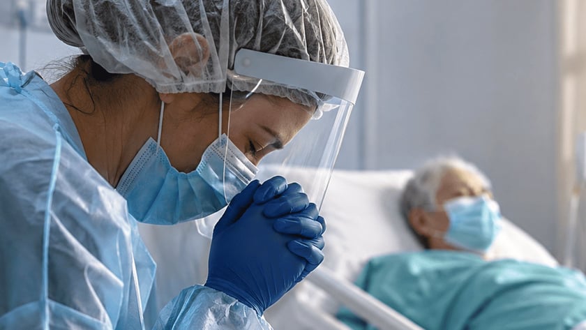 Profissional de saúde com equipamento de proteção junta as mãos ao lado de paciente deitado