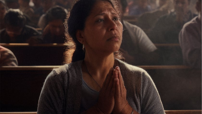 Mulher de cardigã cinza reza de mãos juntas em um banco de igreja lotada.