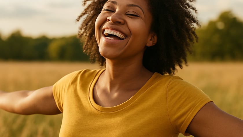 Mulher de cabelo cacheado e camiseta amarela sorri de braços abertos em um campo.