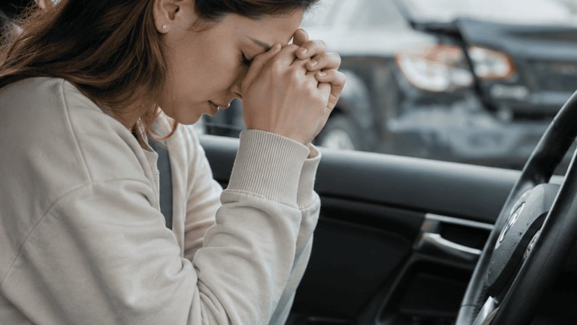 Mulher sentada ao volante de um carro, com o rosto apoiado nas mãos unidas.