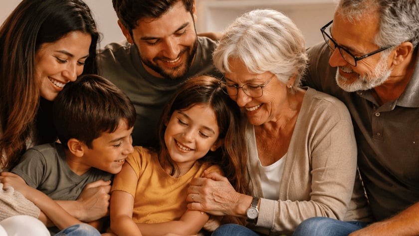 Família de três gerações sentada junta em um sofá, sorrindo e abraçando duas crianças na sala.