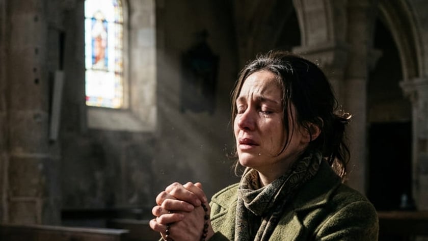 Mulher de casaco verde ajoelhada em uma igreja, com um terço nas mãos, chorando.