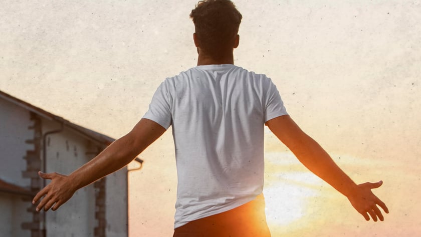 Homem de costas, com os braços abertos, olha o pôr do sol perto de uma casa.