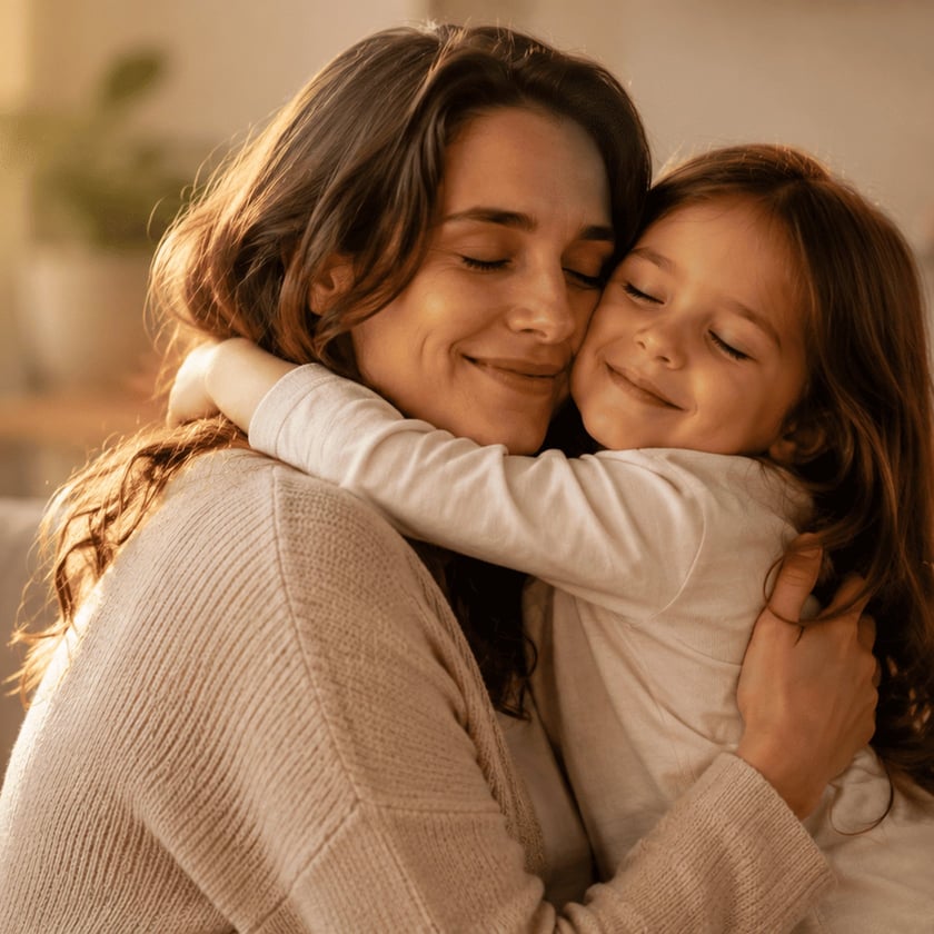 Mulher de cabelos castanhos abraça uma menina sorridente, ambas de olhos fechados, dentro de casa.