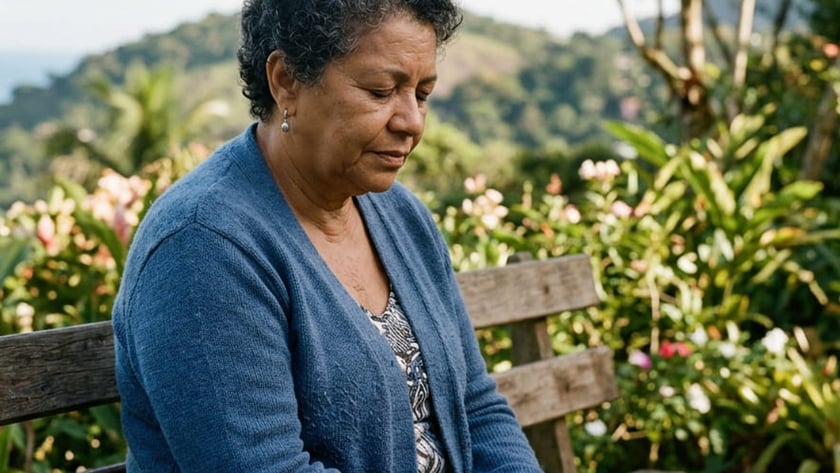 Mulher de cabelo curto e casaco azul sentada num banco de madeira, de mãos unidas, em jardim.