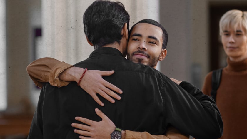 Dois homens se abraçam em uma igreja, enquanto um jovem loiro os observa ao fundo.