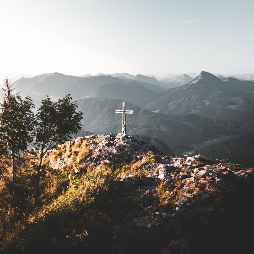 Cruz de metal no topo rochoso de uma montanha, com cadeias de montanhas ao fundo.