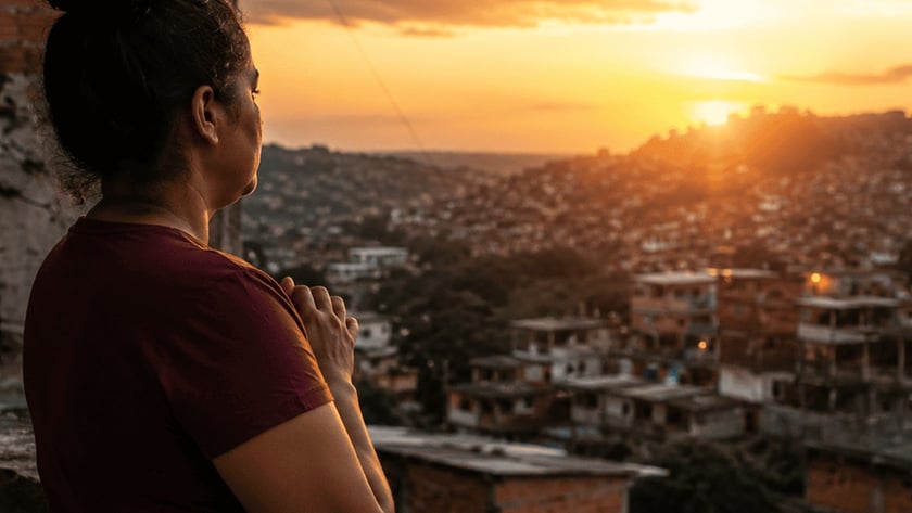 Mulher de camiseta vinho, de costas, observa o pôr do sol sobre uma comunidade em colinas.
