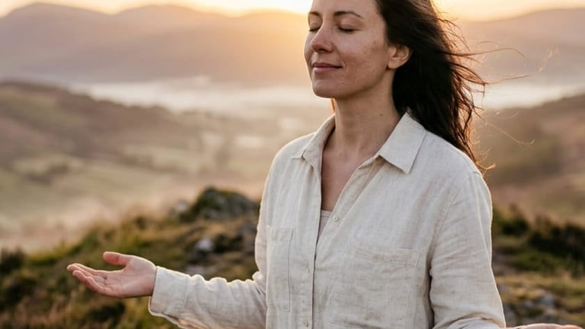 Mulher de olhos fechados e braços abertos em um morro com montanhas ao fundo.