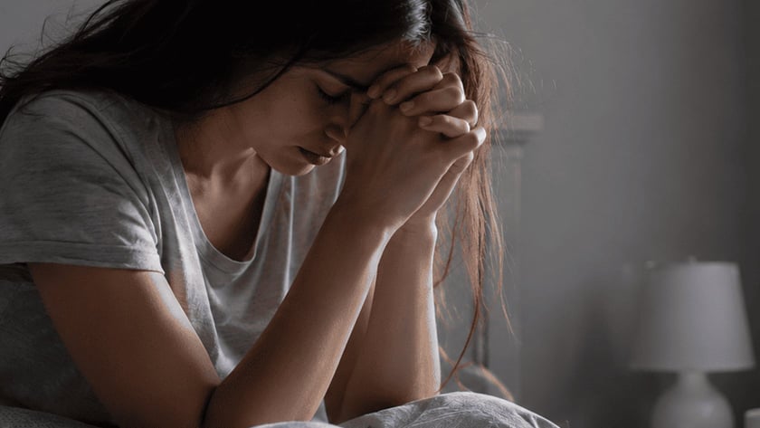 Mulher de cabelos longos sentada na cama, com a cabeça apoiada nas mãos unidas.