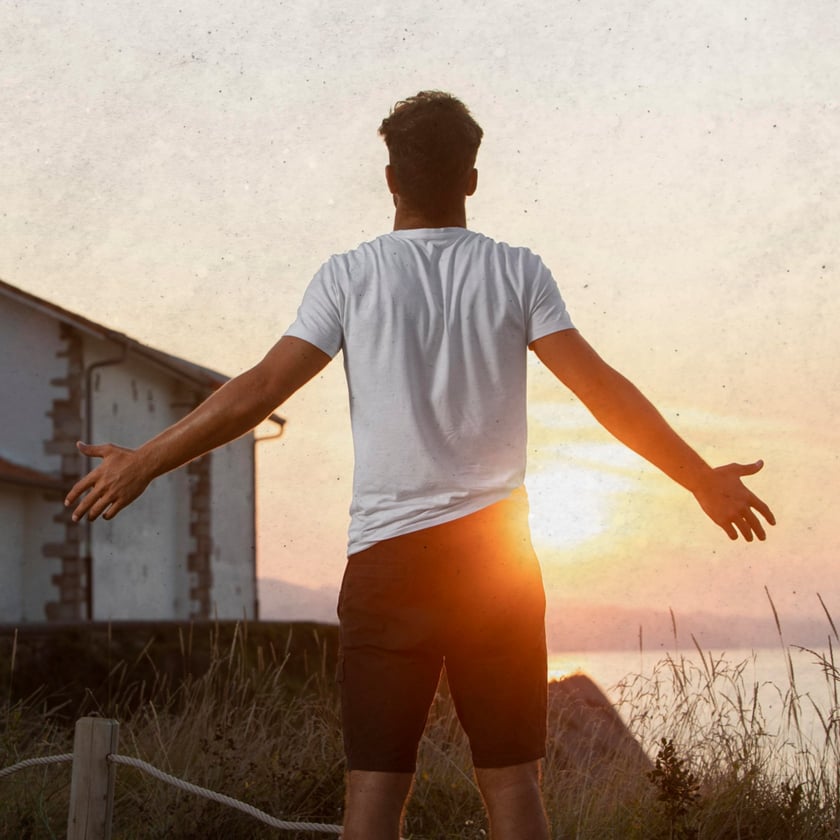 Homem de costas, com os braços abertos, olha o pôr do sol perto de uma casa.