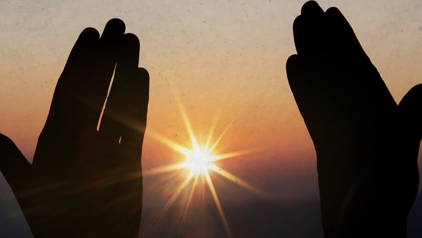 Duas mãos erguidas em silhueta enquadram o sol brilhando no horizonte ao entardecer.