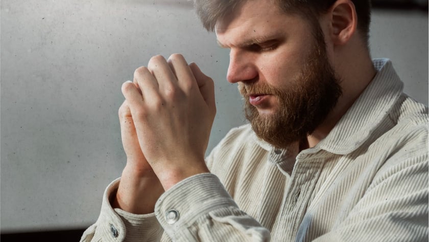 Homem barbudo de camisa bege com as mãos unidas, de olhos fechados, diante de um livro.