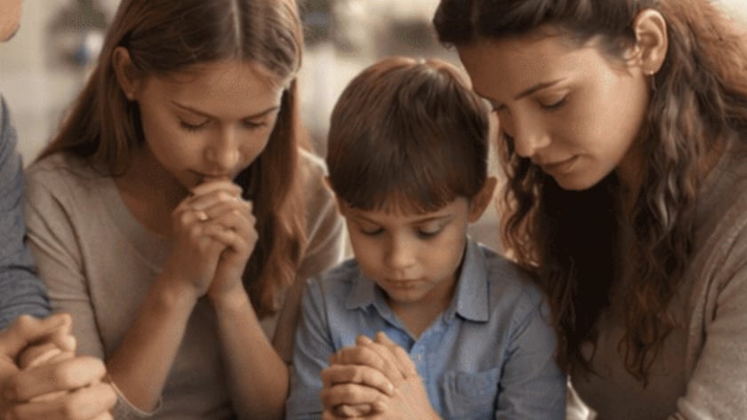 Mulher, menina e menino sentados juntos, de olhos fechados e mãos unidas, rezando.