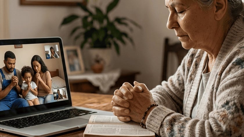 Idosa de mãos postas sobre uma Bíblia reza diante de um notebook com videochamada de família.
