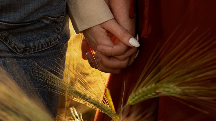Duas pessoas de mãos dadas em um campo de trigo, uma de jeans e outra de vestido vermelho.